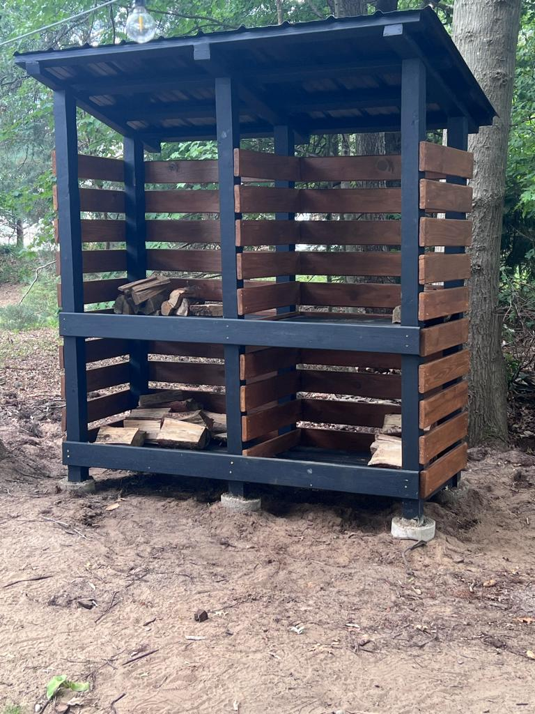Customer-built tiered firewood shed with dark painted posts and stained reclaimed deck boards, standing in a wooded garden