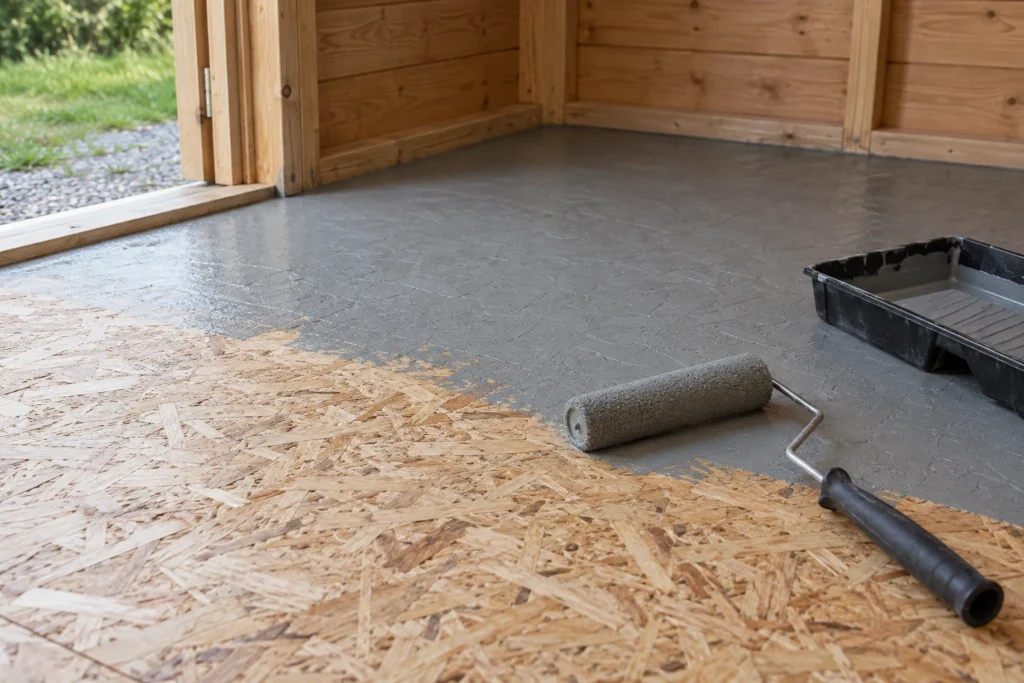 A roller applying a thin grey floor coating to a dry OSB shed floor